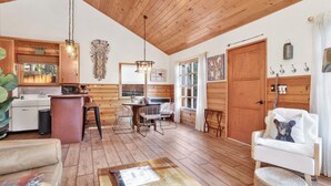 A cozy cabin interior with wooden walls and ceiling, a dining area with a table and chairs, a kitchen with a sink and stove, and a pet-friendly setup with a dog bed and a cat tree.