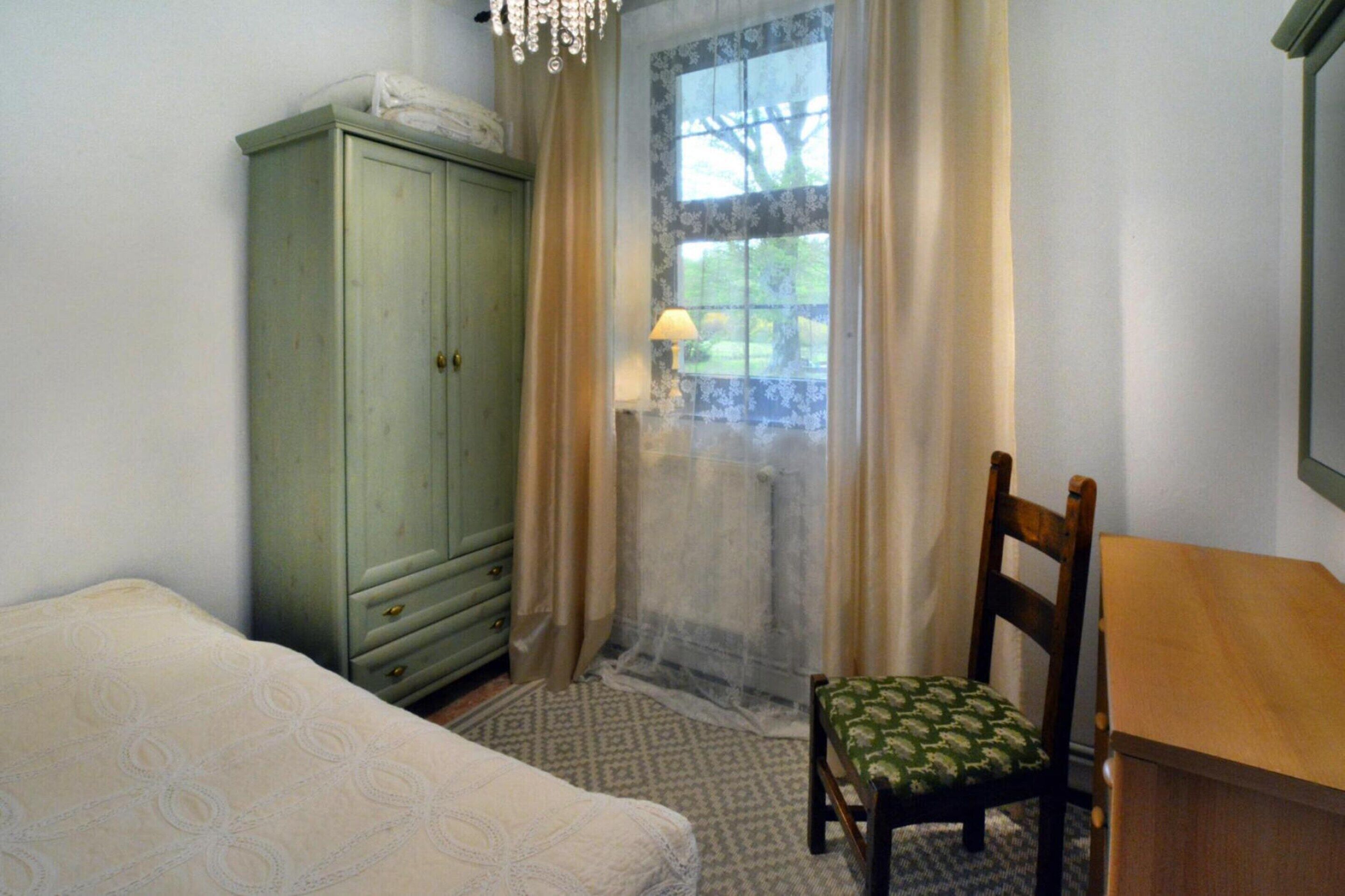 Une chambre à coucher avec un lit, une chaise en bois, un bureau en bois, une armoire verte et une fenêtre avec des rideaux.