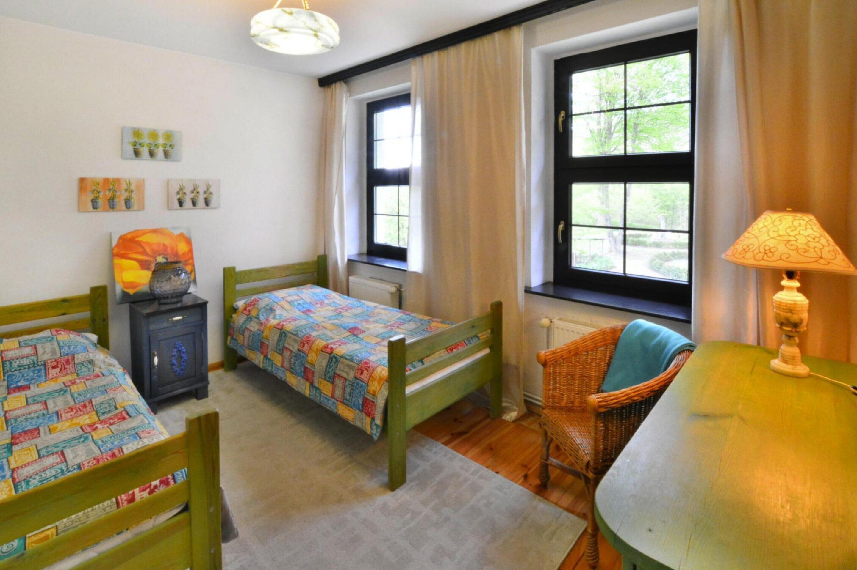 Un dormitorio con dos camas de madera, una silla de mimbre, una mesa con lámpara y una ventana con cortinas.
