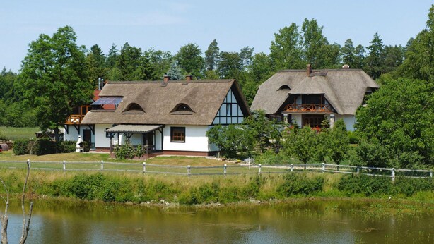 Een traditioneel huis met rieten dak en een witte gevel, gelegen aan een rustig meer, omringd door weelderige begroeiing.