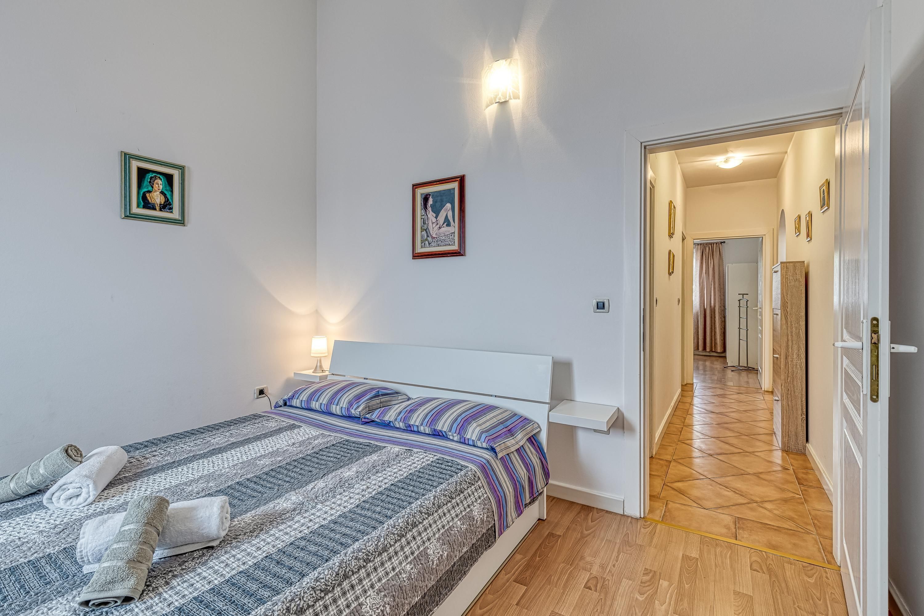 A bedroom with a bed, two wall-mounted lights, a framed picture, and a hallway leading to a bathroom.