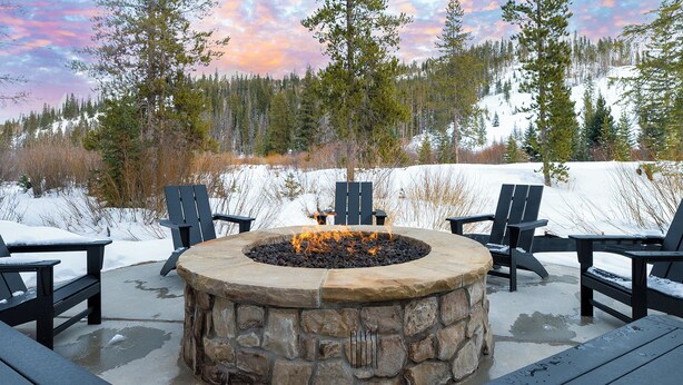 A stone fire pit surrounded by black Adirondack chairs, set against a snowy landscape with trees and a clear sky.