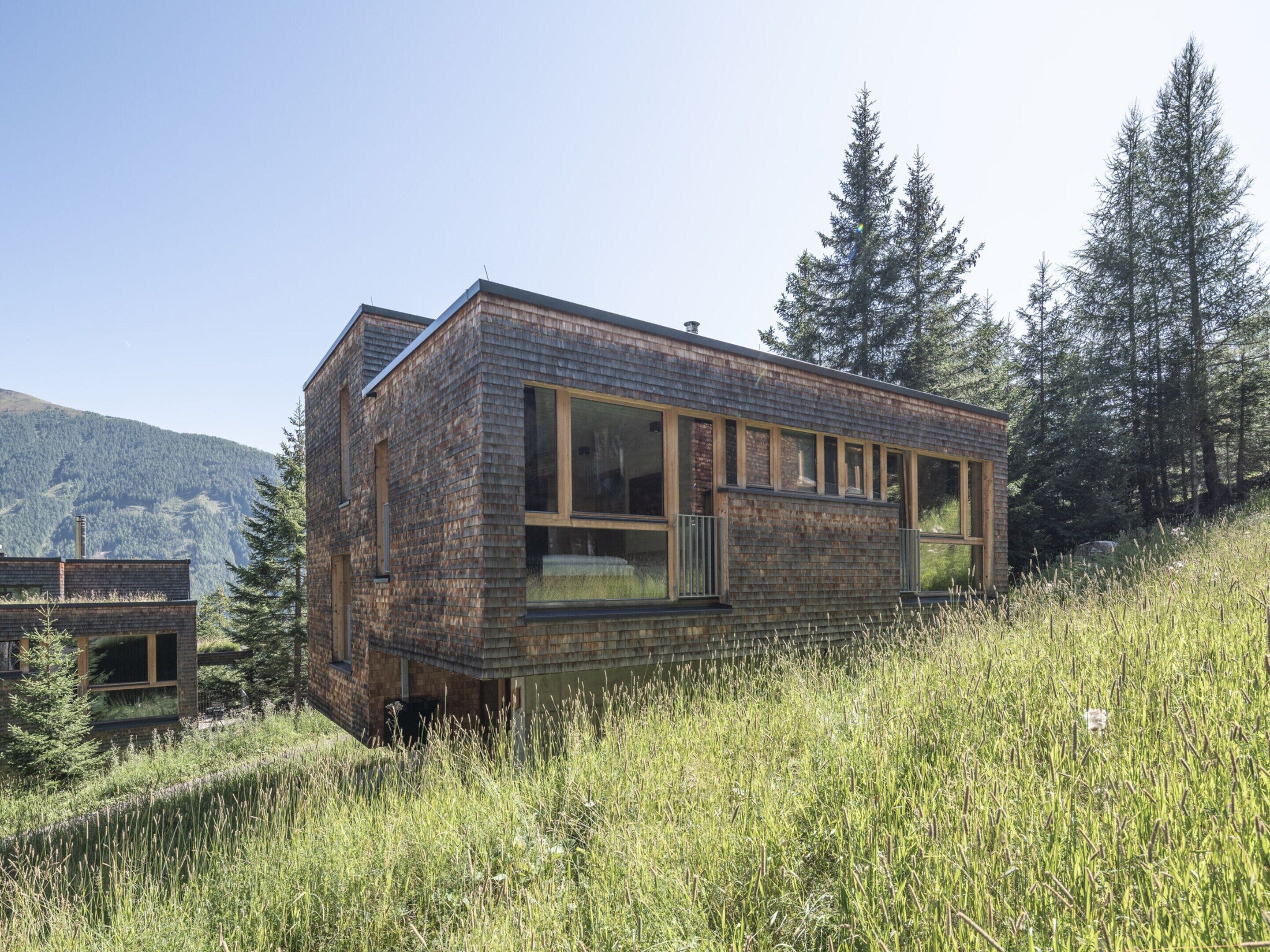 Een modern, rustiek vakantiehuisje met grote ramen en een schuin dak, omgeven door hoog gras en bomen.