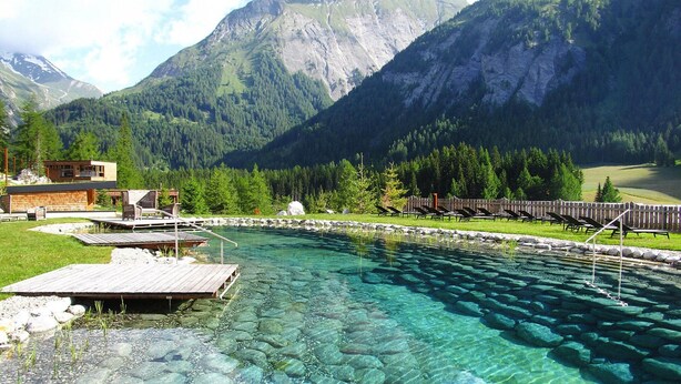 A mountain resort with a clear pool, wooden deck, and lounge chairs. - Chalet Resort Near Lake (Kals am Grossglockner)