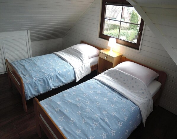 Un dormitorio pequeño y acogedor con dos camas individuales, cada una con una colcha azul estampada con estrellas y almohadas blancas. Hay una mesita de noche de madera con una lámpara entre las camas, y una ventana sobre la cama derecha que ofrece vistas al exterior.