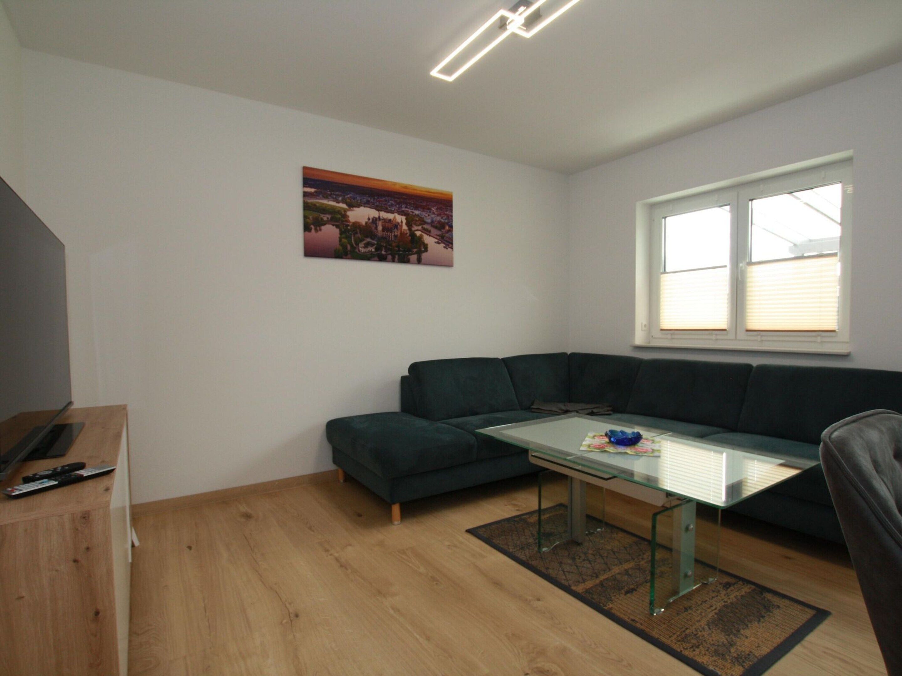 A modern living room with a dark green sectional sofa, a glass coffee table, and a wooden floor. There is a window with blinds, a painting on the wall, and a small table with a blue bowl.