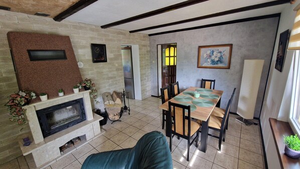 A living room with a fireplace, dining table, and chairs. There is a TV mounted on the wall, a window with blinds, and a door leading to another room. - Holiday Home in Kopice by Stepnica for 5 Persons (Stepnica)