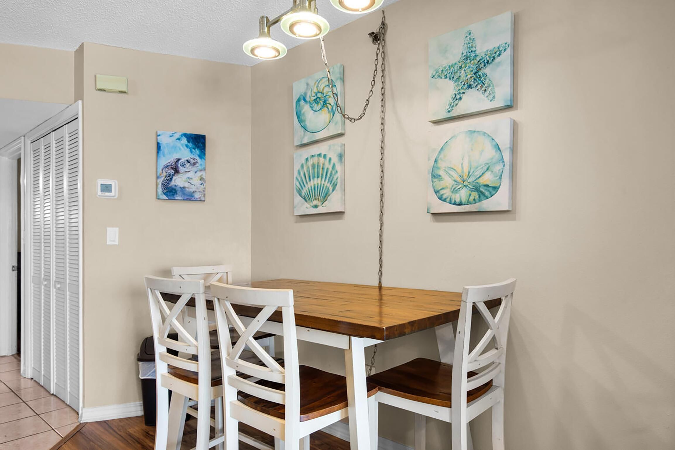 A dining area with a wooden table and white chairs, a ceiling light fixture, and wall-mounted artwork.