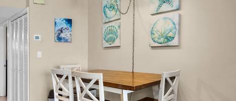 A dining area with a wooden table and white chairs, a ceiling light fixture, and wall-mounted artwork.