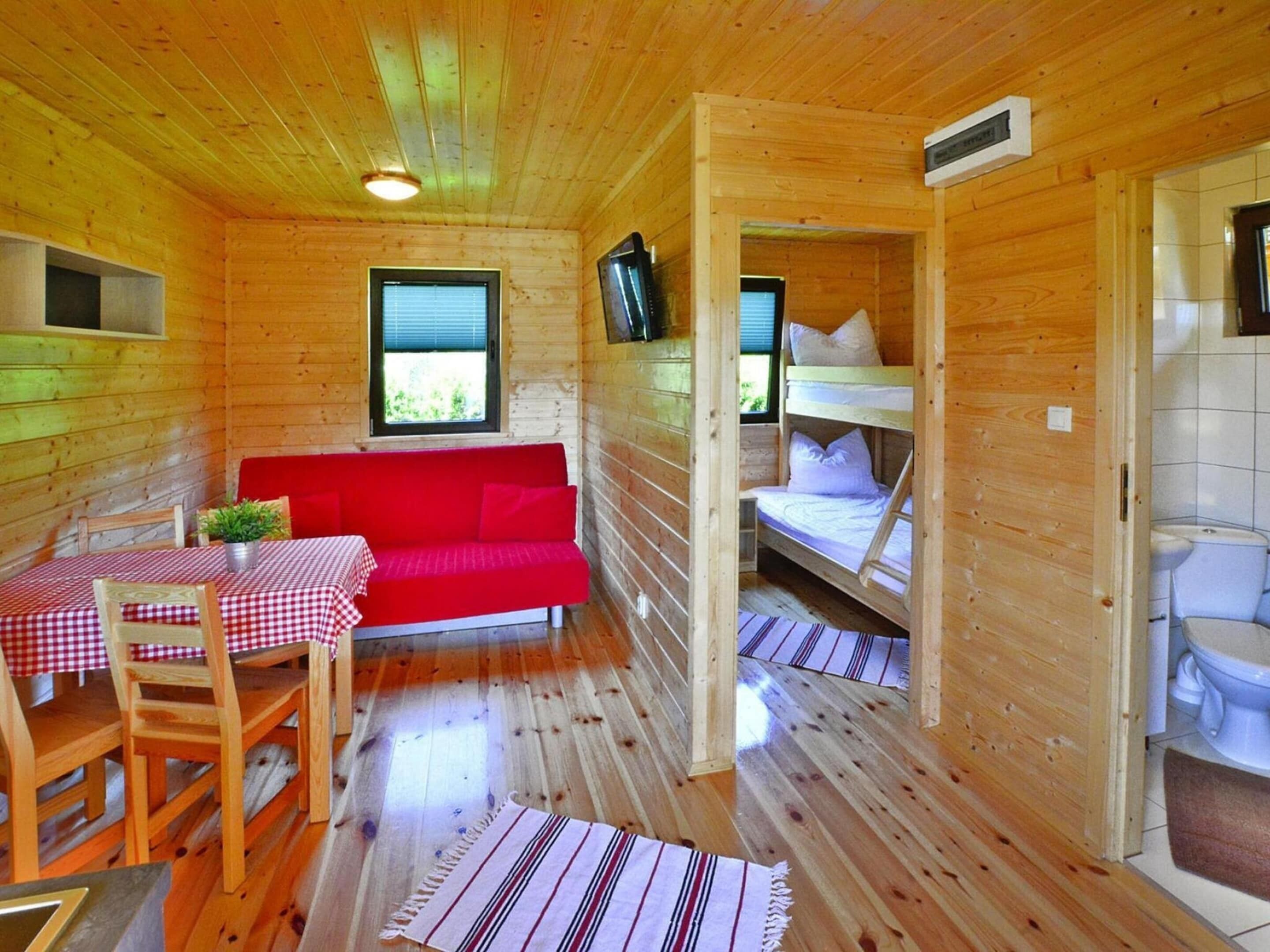 A cabin with a red sofa, a table with checkered tablecloth, a bunk bed, and a small kitchenette.