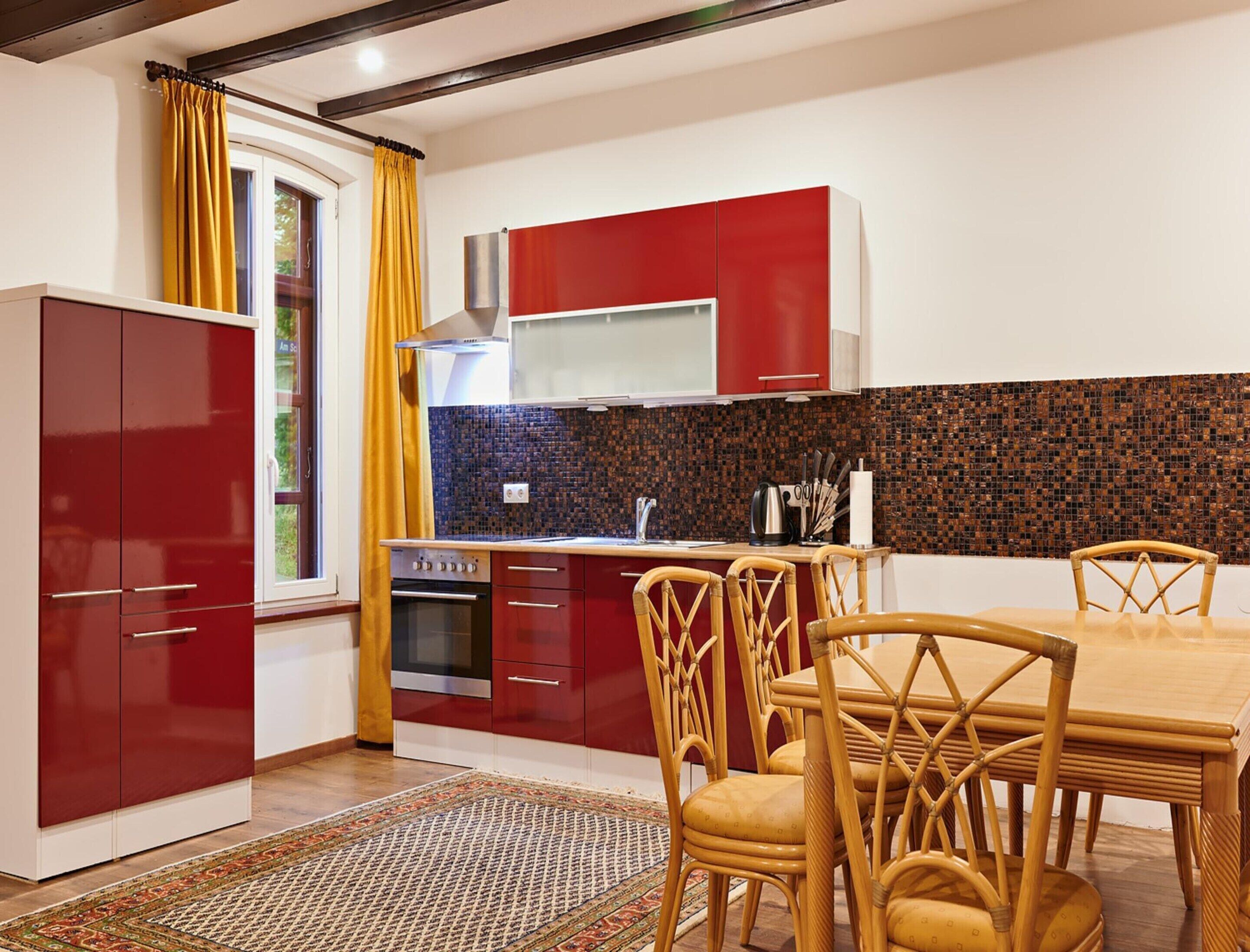 A modern kitchen with red cabinets, a tiled backsplash, and a dining area with wicker chairs and a wooden table.