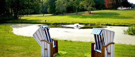 Two wicker lounge chairs with striped cushions in a grassy park area with a pond and trees in the background.