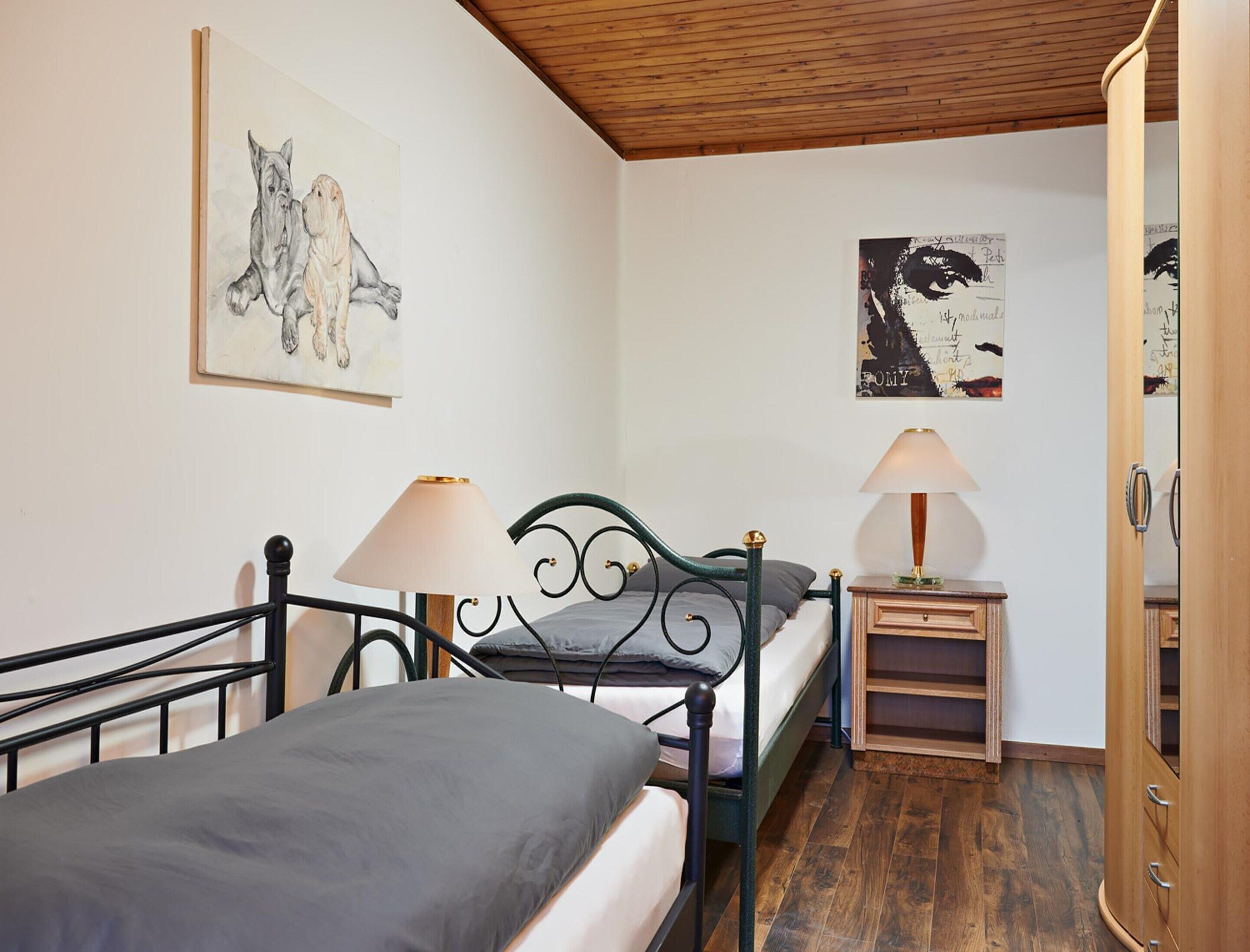 A bedroom with a bed, two bedside tables with lamps, a wooden wardrobe, and two wall-mounted artworks.