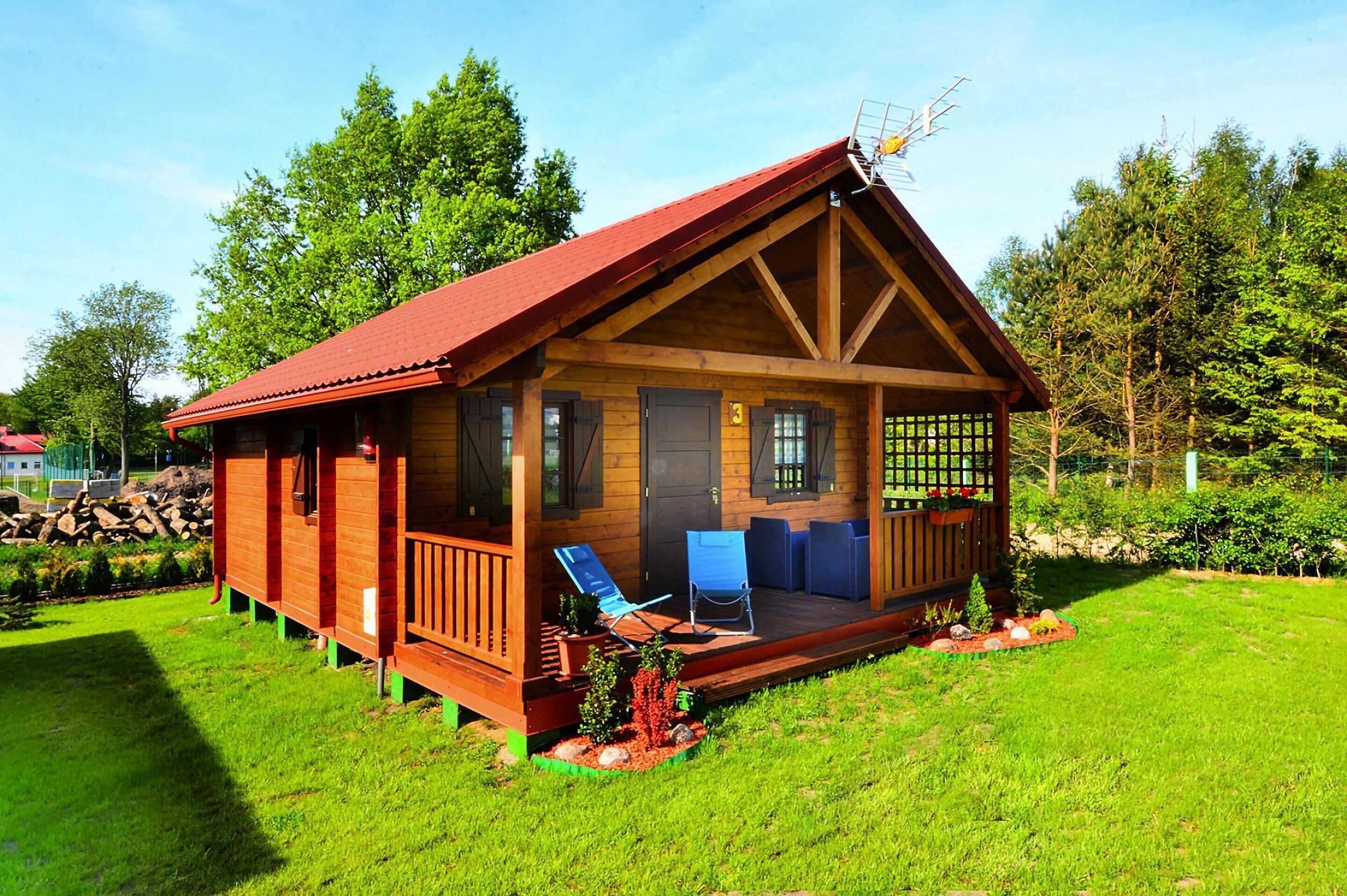 A wooden cabin with a red roof, a porch with blue chairs, and a small garden area.