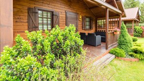 Une cabane en bois dotée d’un porche, de deux fenêtres avec volets et d’un petit jardin agrémenté d’arbustes et d’une plante en pot.