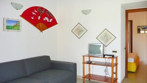Un soggiorno con un divano grigio, un mobile TV in legno con televisore e una ventola rossa con uccelli bianchi.