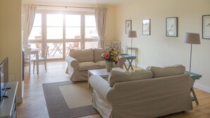 Ein Wohnzimmer mit zwei Sofas, einem Couchtisch und einem Fernseher. Ein Essbereich mit Stühlen und einem Tisch ist durch die Glastüren sichtbar.