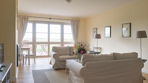Ein geräumiges Wohnzimmer mit zwei Sofas, einem Couchtisch und einem Fernseher. Es gibt ein großes Fenster mit Vorhängen, einen Essbereich mit Tisch und Stühlen sowie einen kleinen Beistelltisch mit Lampe und einer Blumenvase.