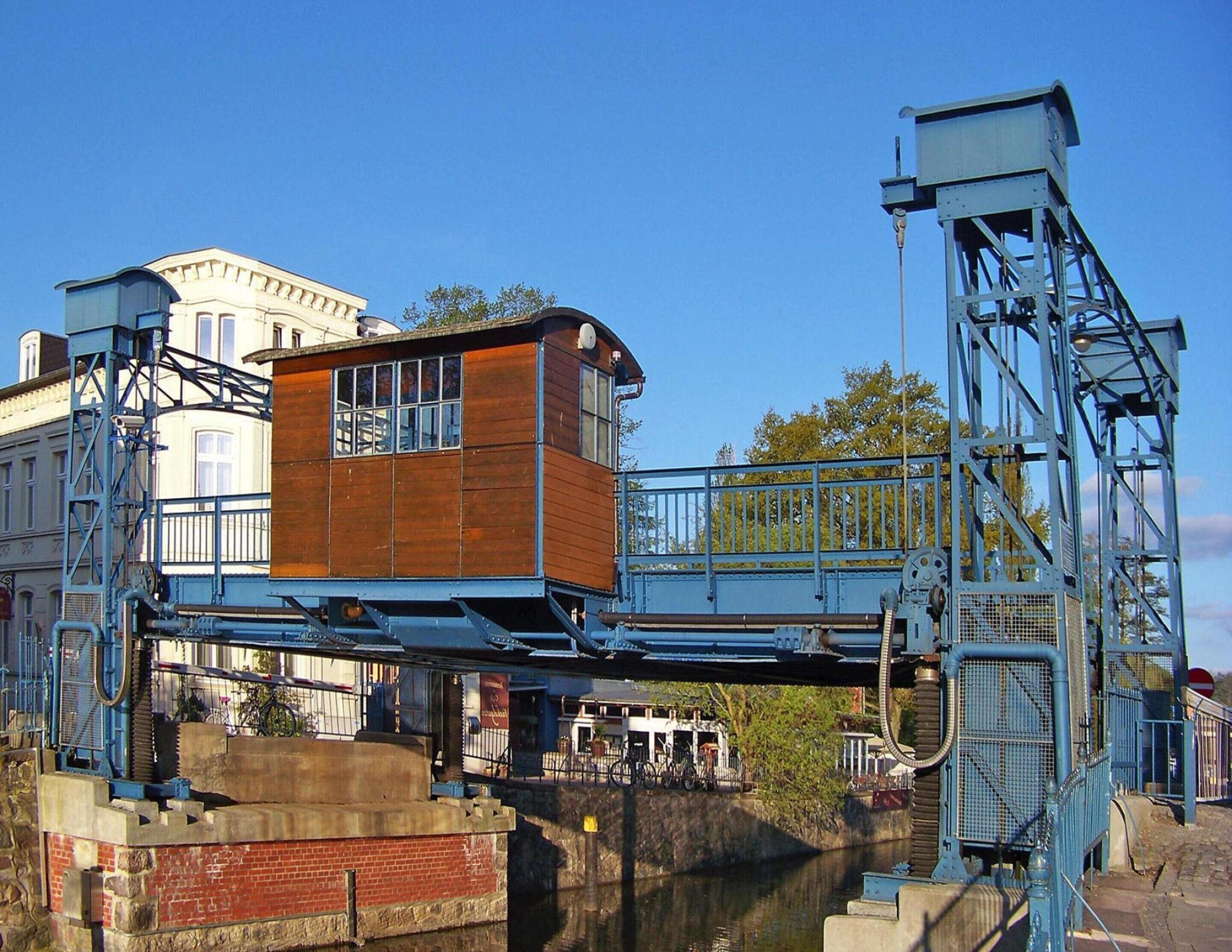 A wooden building with a metal bridge structure over a canal, surrounded by buildings and trees.