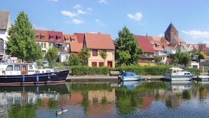 A calm river with boats docked on the left, surrounded by traditional European-style buildings with red roofs and a church tower. - Appartement Hafenflair am Plauer See (Plau am See)