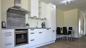A modern kitchen with white cabinetry, a stainless steel range hood, and a built-in oven. There is a dining area with a table and chairs. - Appartement Hafenflair am Plauer See (Plau am See)