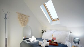 Ein Schlafzimmer mit schrägen Wänden, einem Bett mit dunkelblauer Steudecke, weißen Kissen, einem Nachttisch mit Flasche und Gläsern sowie einer Stehlampe.
