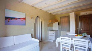 A living room with a white sofa, a dining table with chairs, a kitchen area with a stove, and a painting on the wall. - Golfo Aranci Stay at Baia de Bahas (Golfo Aranci)