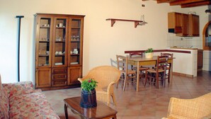 Un comedor con una mesa de madera, sillas y una planta en maceta. Hay un sofá de mimbre y un armario con puertas de vidrio.