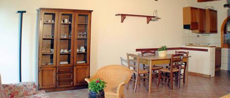 Un comedor con una mesa de madera, sillas y una planta en maceta. Hay un sofá de mimbre y un armario con puertas de vidrio.