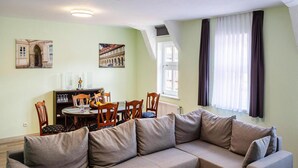 Un salon moderne avec un canapé d’angle, une table à manger et des chaises. On y trouve deux tableaux accrochés aux murs, une fenêtre avec des rideaux et un radiateur.