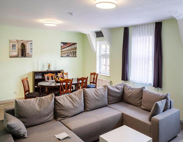 Uma sala de estar moderna com sofá modular, mesa de jantar e cadeiras. Há duas pinturas nas paredes, uma janela com cortinas e um radiador.