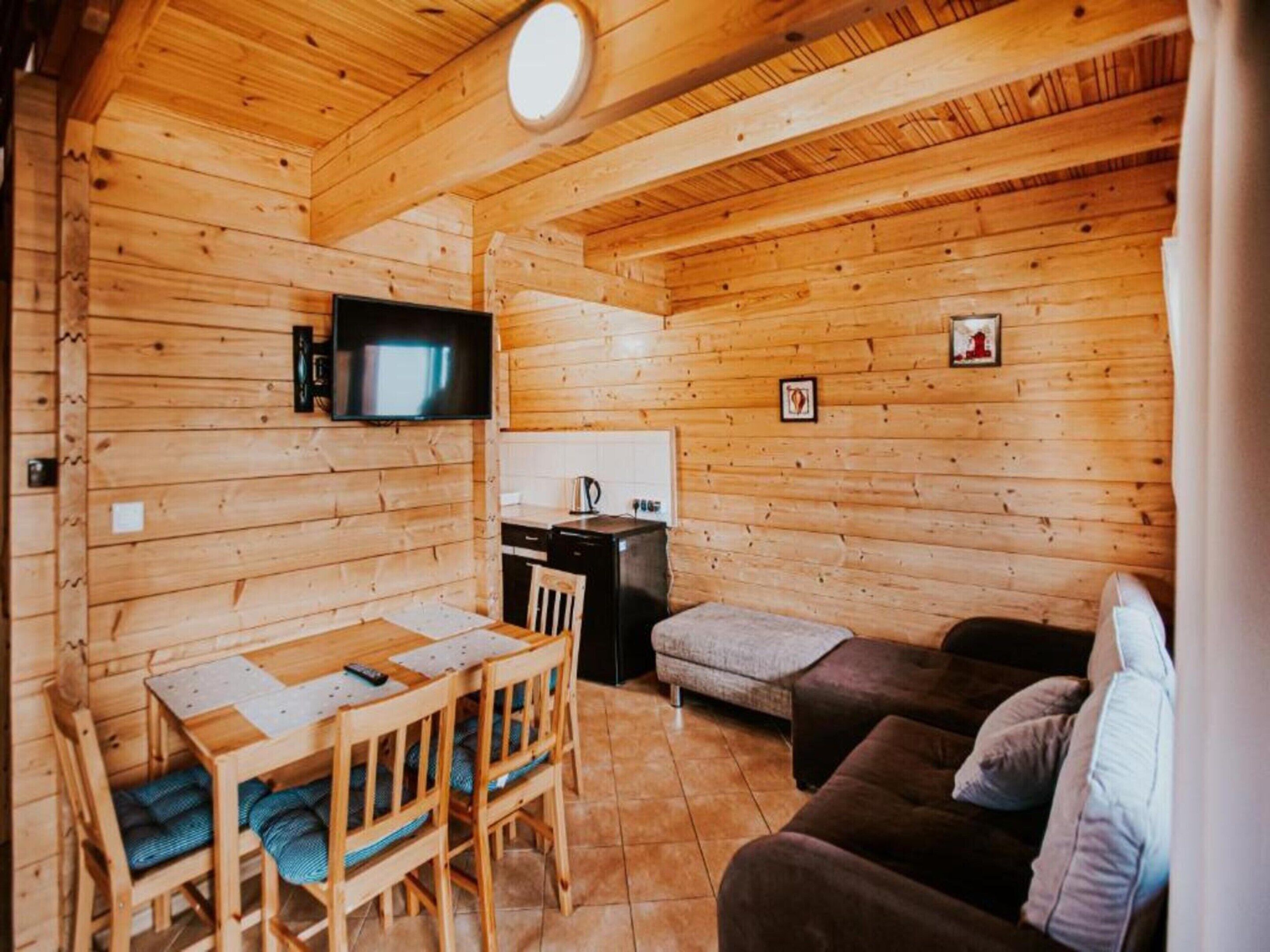 A wooden cabin interior with a dining table, chairs, a sofa, a television, and a kitchen area.