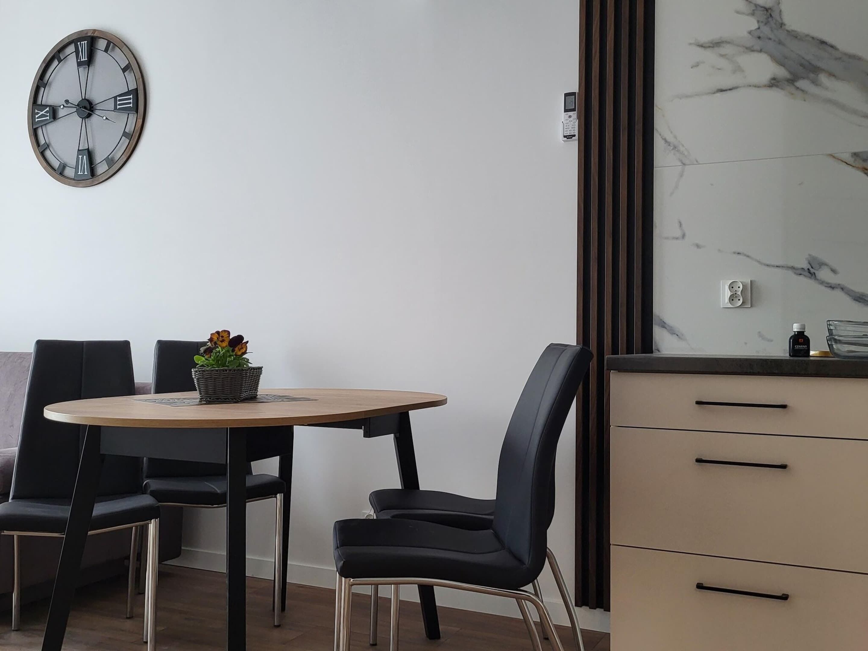 A modern dining area with a round table, black chairs, a clock, and a marble backsplash.