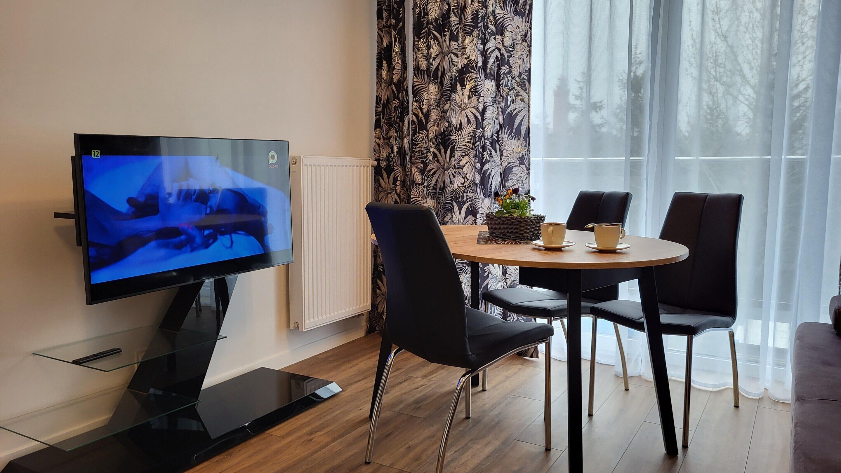 Una sala moderna con un televisor de pantalla plana, una mesa redonda de madera, sillas negras y un ventanal con cortinas traslúcidas.