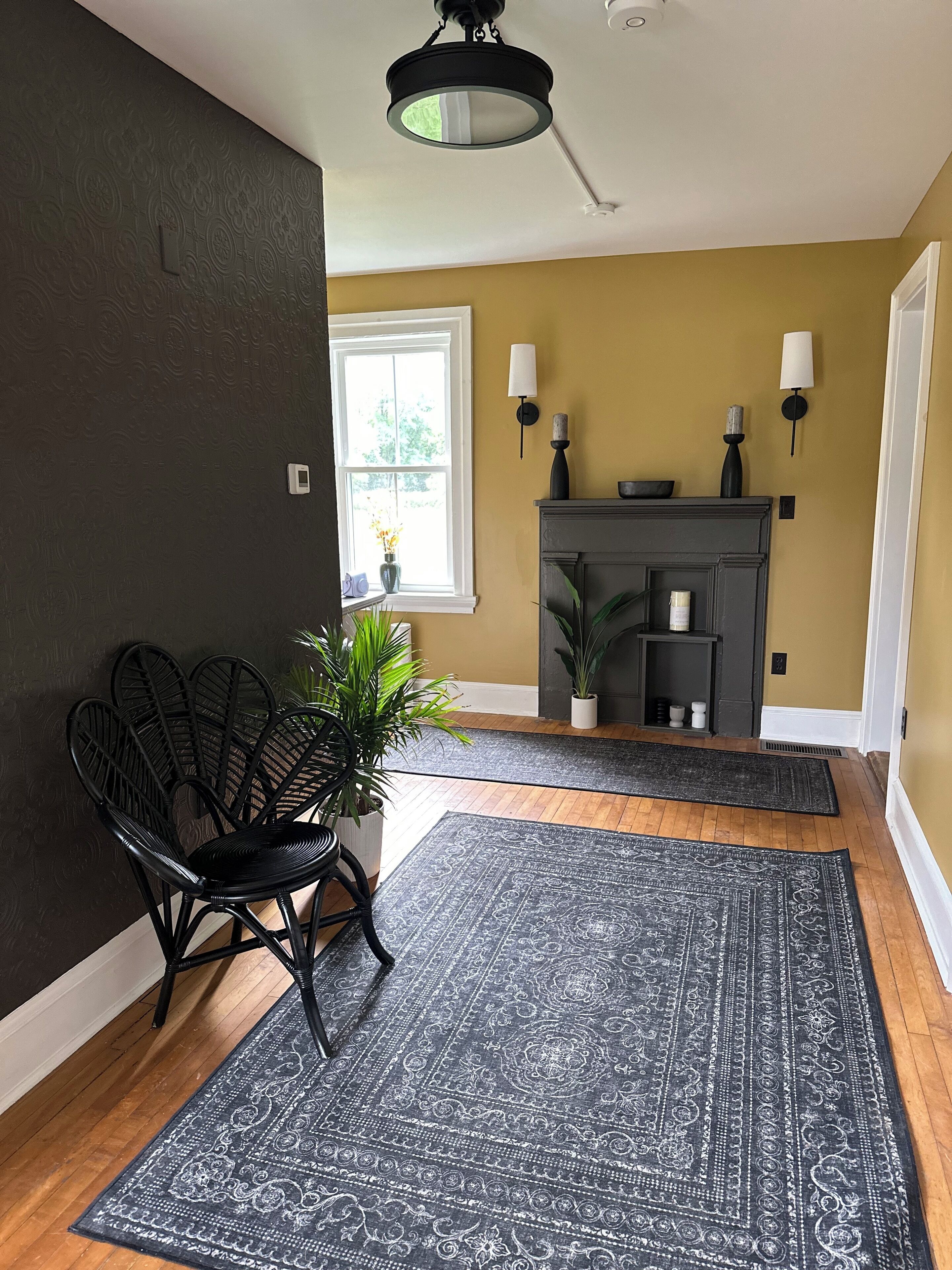 Una habitación con una pared oscura, una ventana, una chimenea, una silla negra y una alfombra estampada.