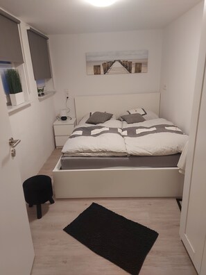 A small, neatly organized bedroom with a bed, bedside tables, a nightstand, a window with blinds, a wall-mounted picture, and a black rug on the floor. - Quiet central granny apartment (Bad Hersfeld)