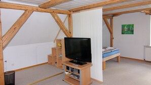 A room with a sloped ceiling, wooden beams, a television on a wooden stand, a bed with a blue mattress, and a small shelf with items.