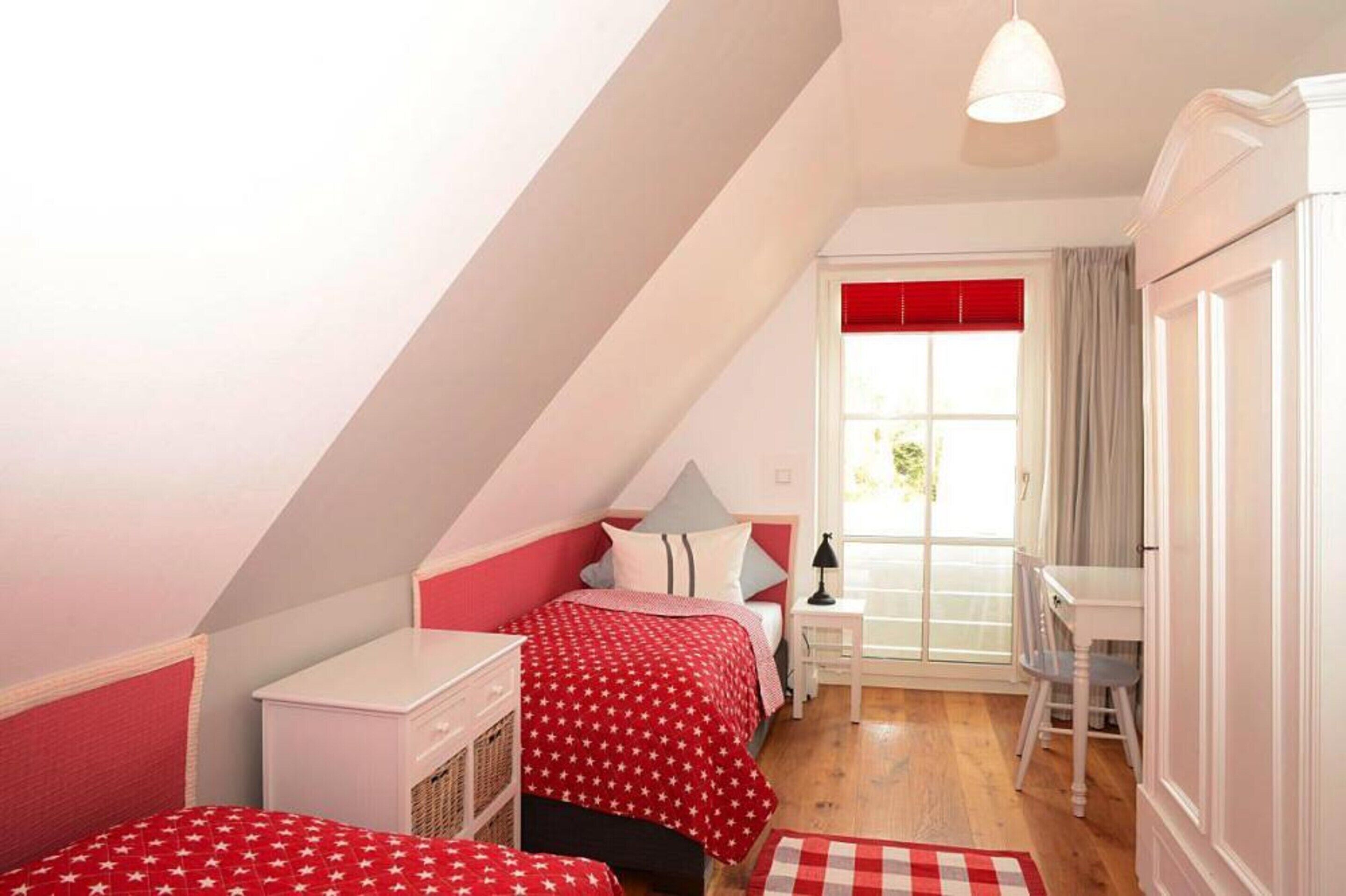 Ein Schlafzimmer mit schrägen Wänden, einem Bett mit rot-weißer Bettwäsche, einem kleinen Schreibtisch und einem Fenster mit Vorhängen.
