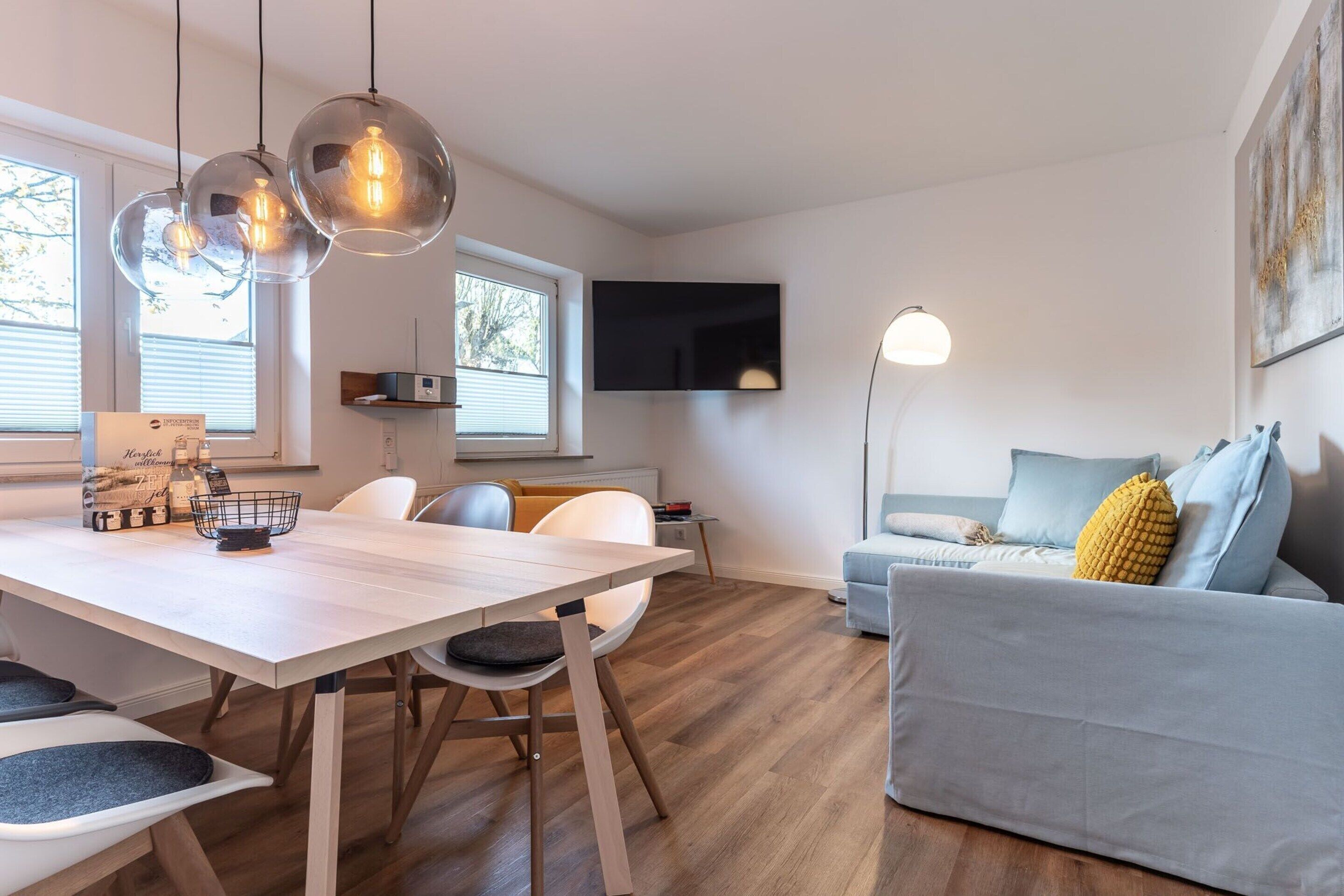 A modern living room with a dining area, a flat-screen TV mounted on the wall, and a sofa with cushions.
