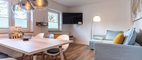 A modern living room with a dining area, a flat-screen TV mounted on the wall, and a sofa with cushions.