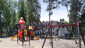 A playground with swings and slides, surrounded by trees and a building in the background. - Holiday Home in Lukecin Near Baltic Sea (Dziwnów)