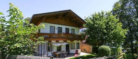 A two-story chalet with a balcony, a dining area with outdoor seating, and a wooden fence in the foreground.