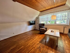Una habitación con suelo de madera, una mesa blanca, un sillón marrón, una lámpara de pie, un televisor montado en la pared y una ventana con cortinas.