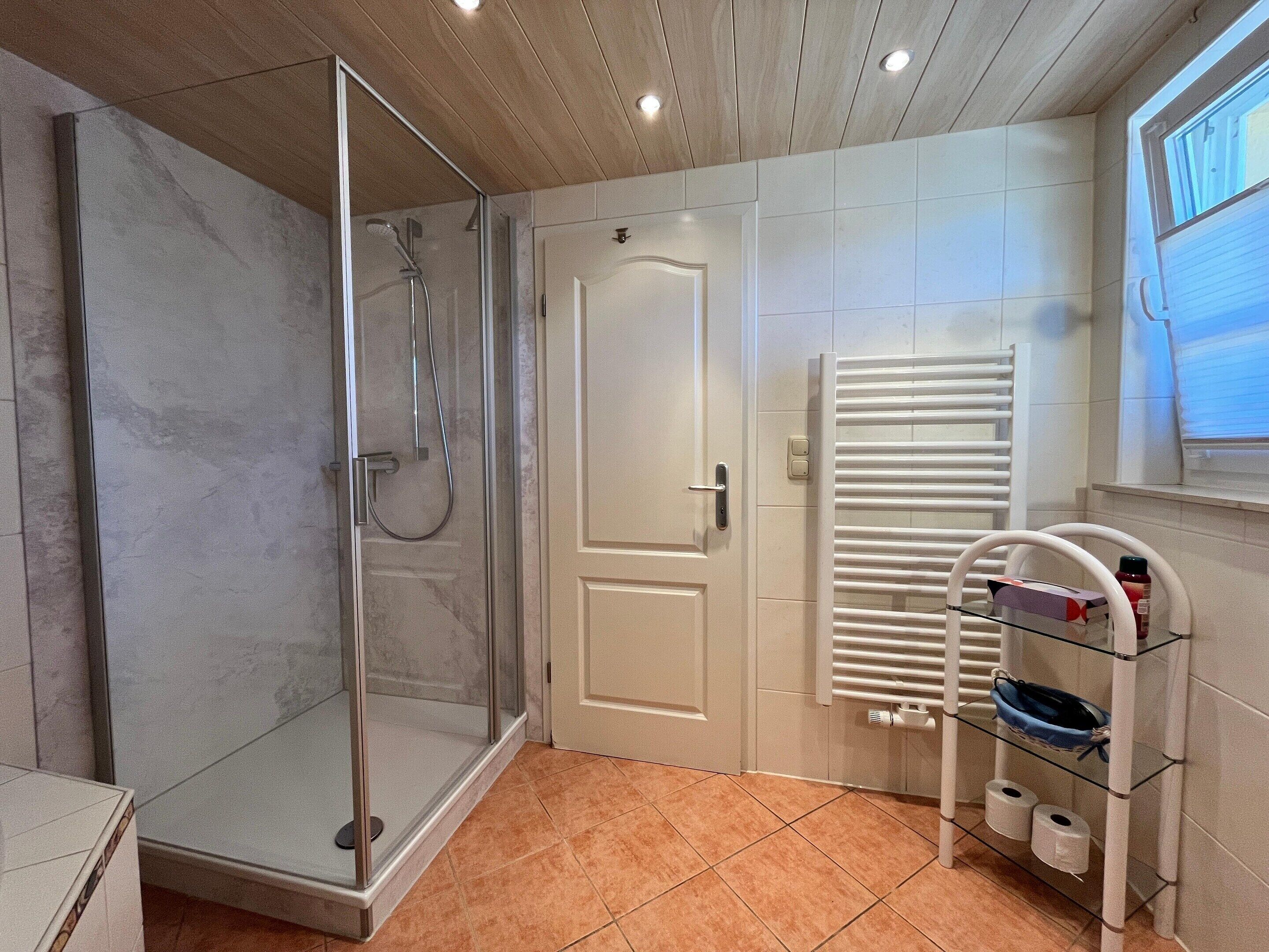 A bathroom with a glass shower enclosure, a white door, a glass shelf with toiletries, and a window.