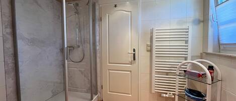A bathroom with a glass shower enclosure, a white door, a glass shelf with toiletries, and a window.