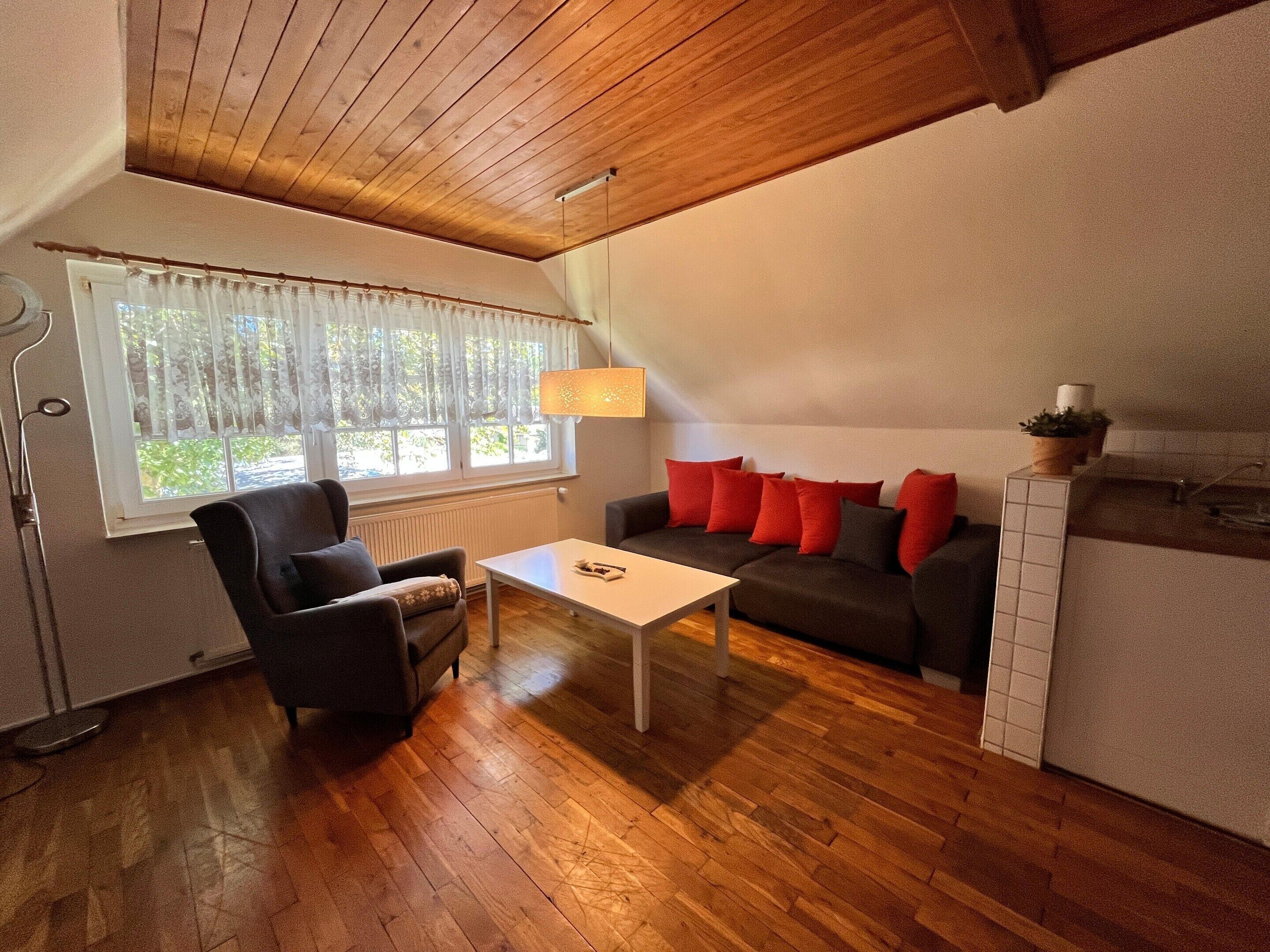 A cozy living room with a sofa, armchair, and coffee table, a window with curtains, and a wooden ceiling.
