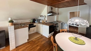 A kitchen with wooden cabinets, a white countertop, a sink, a stove, and a dining area with a table and chairs. - Ferienwohnung, Wieck am Darss (Wieck auf dem Darss)