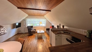 A compact, modern apartment with a sloped ceiling, a dining area, a kitchen, and a large window providing natural light. - Ferienwohnung, Wieck am Darss (Wieck auf dem Darss)