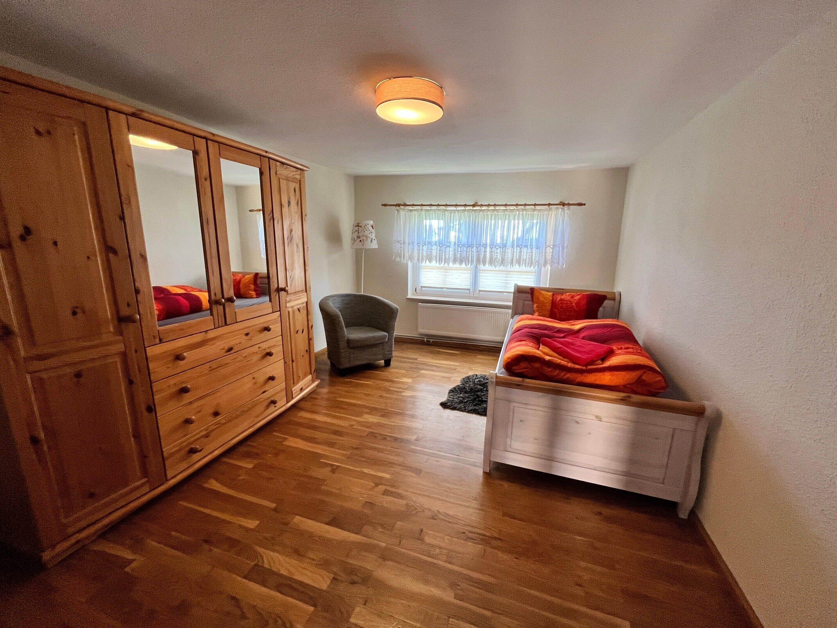 A bedroom with a wooden wardrobe, a single bed with a red blanket, a chair, and a window with curtains.