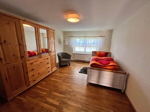 Un dormitorio con un armario de madera, una cama individual con una manta roja, una silla y una ventana con cortinas.